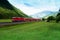 Red train crossing green valley near Alps