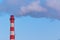 Red and white chimney of a factory, from which gray smoke comes out against a blue sky on a frosty winter day