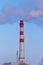 Red and white chimney of a factory, from which gray smoke comes out against a blue sky on a sunny frosty day