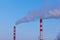 Red and white chimney of a factory, from which gray smoke comes out against a blue sky on a sunny winter day