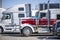 Red and white classic big rig semi truck with low profile cab and high exhaust pipes standing in row with another semi trucks and