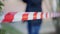 Red and white police tape outdoors with blurred unrecognizable criminalist walking at background. Crime scene with woman