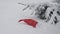 Red and white Santa Claus hat is lying on a snow near spruce outdoor in calm daytime, snow is falling from top