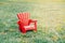 Red wooden plastic muskoka Adirondack chair standing on green grass among yellow dandelion flowers in park outside