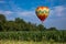 Red, Yellow, Orange, Green, and White Teardrop Shaped Hot Air Balloon Over Corn Field on Sunny Day with Trees and Cloudy Blue Sky