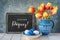 Red-yellow tulips in blue ceramic pitcher with Easter eggs and a blackboard on gray background. Text