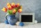 Red-yellow tulips in blue ceramic pitcher with Easter eggs and a blackboard, text space