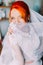 Redhair girl in a wedding dress covers face by veil. Bright appearance, studio interior background