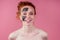 Redhaired ginger teenager girl with black clay mask on her pretty face on pink studio background