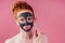 Redhaired ginger teenager girl with black clay mask on her pretty face on pink studio background