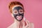 Redhaired ginger teenager girl with black clay mask on her pretty face on pink studio background