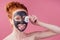 Redhaired ginger teenager girl with black clay mask on her pretty face on pink studio background .