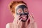 Redhaired ginger teenager girl with black clay mask on her pretty face on pink studio background