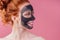 Redhaired ginger teenager girl with black clay mask on her pretty face on pink studio background