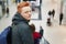 A redhead freckled stylish boy standing back in shopping mall wearing fashionable jacket with hood and holding rucksack on his bac