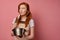 A redhead girl in a apron and a T-shirt stands on a pink background, with a bowl and a whisk in hands and looks scared