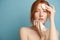 Redhead girl with clean radiant skin, without makeup, wipes her face with a napkin and cotton pad, standing on a blue