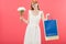 Redhead girl in glasses and white dress holding bouquet of flowers and shopping bags isolated on pink