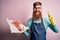 Redhead Irish butcher man with beard holding raw beef steak over pink background surprised with an idea or question pointing