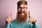 Redhead Irish man with beard holding beauty razor for shaving and skin care over pink background surprised with an idea or