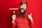 Redhead man with long beard holding fork with pork sausage pointing thumb up to the side smiling happy with open mouth