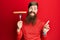 Redhead man with long beard holding fork with pork sausage smiling happy pointing with hand and finger to the side