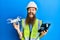 Redhead man with long beard wearing safety hard using drone with remote control smiling and laughing hard out loud because funny