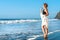 Redhead tender girl stand barefoot in the surf white foam on the beach. Woman in a white dress on the coast of an ocean