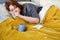 Redhead woman coughs in hand while lying on bed with cup of tea and medical mask