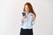 Redhead woman making online order, holding plastic credit card and looking at smartphone screen surprised, white