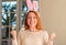 Redhead woman wearing easter rabbit ears at home screaming proud and celebrating victory and success very excited, cheering