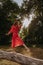 Redhead young woman in red summer country dress balancing and dancing on a dry fallen tree in the middle of forest