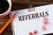 REFERRAL - word on a white sheet on a wooden brown background with a cup of coffee and a pen