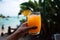 Refreshing beach juice in hands with clear waters and palm trees in the background. Generative AI