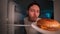 Refrigerator point of view joyful young man opening refrigerator door and taking burger at night with happy face on dark