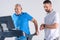 rehabilitation therapist checking time while assisting senior man exercising on treadmill