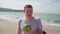 Relaxed adult guy standing and drinking coconut on sandy beach. Handsome man enjoying tropical drink, sipping coconut water