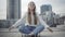 Relaxed Caucasian woman sitting in lotus pose on overcast city square. Portrait of calm smiling young lady enjoying