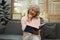 Relaxed elderly woman sitting on comfortable sofa in living room with cup of coffee and reading book