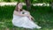 Relaxed girl in white dress sitting on green grass in summer garden. Happy girl teenager relaxing on green lawn in