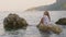 Relaxed girl in white tunic enjoying sea landscape on stone in water. Happy young girl sitting on rock in sea water on