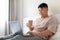 Relaxed korean man drinking coffee and using laptop in bed