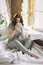 Relaxed positive young brunette woman in homewear sitting on top of unmade bed with cup of tea