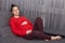 Relaxed pregnant young female rests at couch at home, keeps hands on belly, wears knitted sweater and leggings, looks at camera