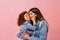 Relaxed preteen girl embracing with mother. Winsome young mom kissing daughter on pink background