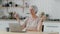 Relaxed pretty elderly grey-haired lady sit at the table in the kitchen with laptop and meditating with closed eyes