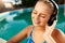 Relaxed smiling woman listening to music in headphones bathing in swimming pool. Blonde girl enjoys favourite song with