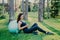 Relaxed sporty young female model takes break after aerobics exercises with fitness ball, sits on green grass, uses mobile phone,