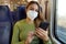 Relaxed woman on train wearing medical face mask using smart phone app. Commuter with protective mask traveling sitting in