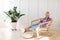 Relaxed woman wearing wireless headphones and using smartphone, sitting in wicker chair at home, free space